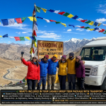 Ladakh group tour package with transport 2026 - group of 6-8 travelers at Khardung La pass 18390 feet posing for group photo, white Tempo Traveller with group tour branding, Ladakhi driver giving thumbs up, colorful jackets, prayer flags, Colors of Ladakh 6 days ₹27,500 per person, Magnificent Ladakh 7 days ₹49,000-52,900, group size pricing 2-3 pax ₹35k-45k, 4-6 pax ₹25k-35k, 7-10 pax ₹20k-28k, 11-15 pax ₹18k-25k