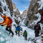 Zanskar Valley Chadar trek package 2026 - group of trekkers walking on frozen Zanskar River with microspikes, ice cliffs and frozen waterfalls on both sides, colorful winter jackets, ice axe, prayer flags in distance, temperature -25°C, Universal Adventures ₹19,999 per person, Himtrek ₹24,500, Himalayan Hikers ₹25,000 + GST, Shikhar Travels ₹34,500, fixed departure dates 13 Jan, 27 Jan, 10 Feb 2026, 9-day itinerary from Leh to Nerak and return, includes heated hotel, camping on ice, -20°C sleeping bags, microspikes, gaiters, ice axes, guides, permits, first aid, oxygen, transport