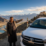 Best cab service in Leh Ladakh 2026 - professional Ladakhi driver with white Toyota Innova SUV at viewpoint overlooking Indus River valley with snow-capped Himalayan mountains