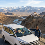Best taxi service in Leh for sightseeing 2026 - professional Ladakhi driver with Toyota Innova SUV at scenic viewpoint overlooking Indus River valley Ladakh Himalayas