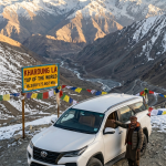 Fortuner for Ladakh trip 2026 - White Toyota Fortuner 4x4 SUV at Khardung La pass 18380 feet world's highest motorable road with prayer flags and Himalayan mountains