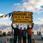 Khardung La pass highest motorable road 2026 - iconic signboard at 18380 feet with happy travelers and prayer flags