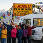 Luxury Ladakh group tour package 2026 - happy group of 12 travelers with white Force Urbania luxury tempo traveller at Khardung La pass 18380 feet