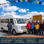 Tempo traveller hire Leh for group 2026 - white Force Urbania at Khardung La pass 18380 feet with happy group of 10-12 travelers posing for photo, Ladakhi driver giving thumbs up, prayer flags, official rates Leh local ₹3,300, Nubra Valley 2 days ₹14,571-17,356, Pangong Tso 2 days ₹16,288, Nubra-Pangong circuit 3 days ₹29,529, Srinagar to Leh 2 days ₹27,831, Tso Moriri 2 days ₹18,655-22,584, vehicle options 9-17 seater, Force Urbania 12-seater most popular, extra charges night halt ₹385, group tour packages 5-14 days ₹22,000-36,500 per person